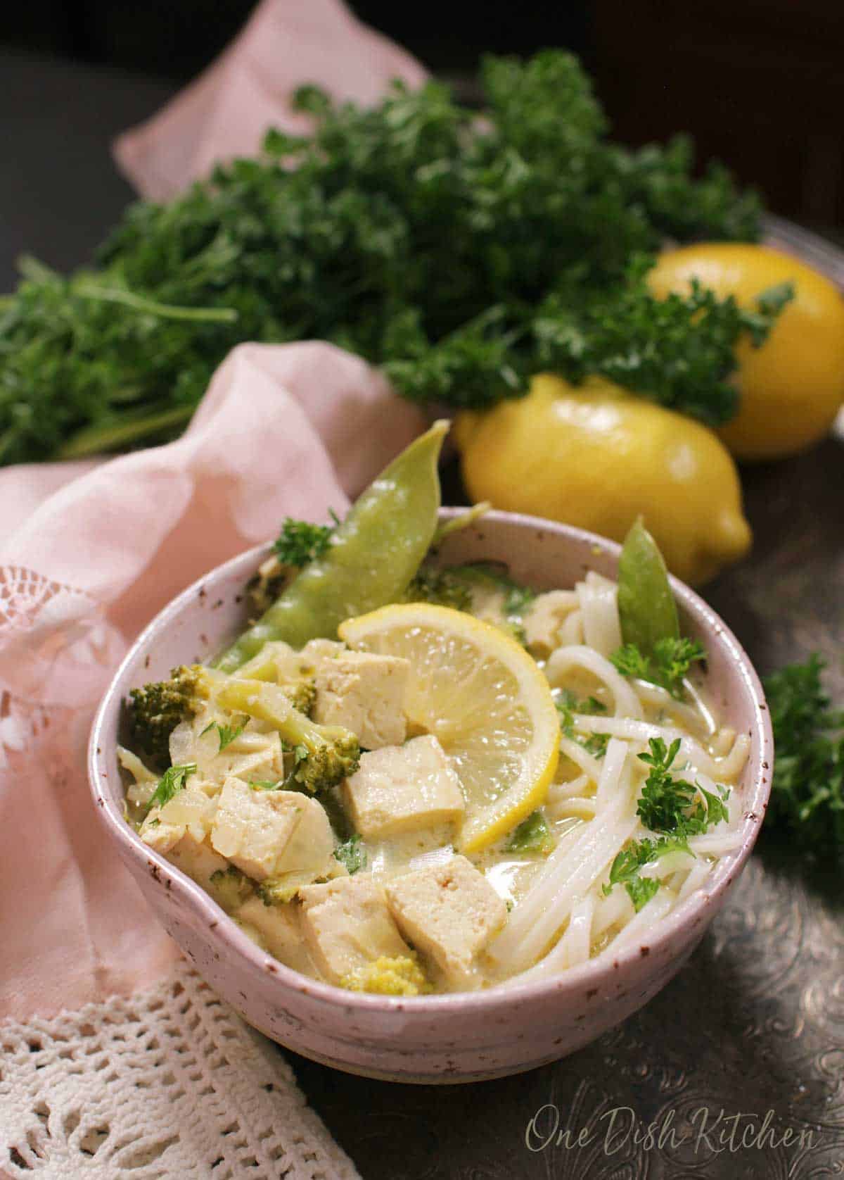 A bowl of green curry with tofu and noodles on a metal tray with a pink cloth napkin, parsley, and two lemons