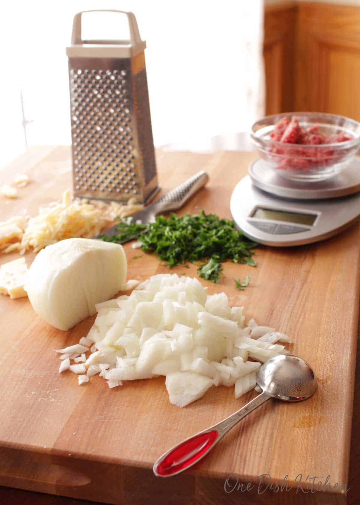 Ingredients for making a meatball on a wooden cutting board- chopped onions, chopped parsley, grated parmesan cheese, and ground beef on a scale