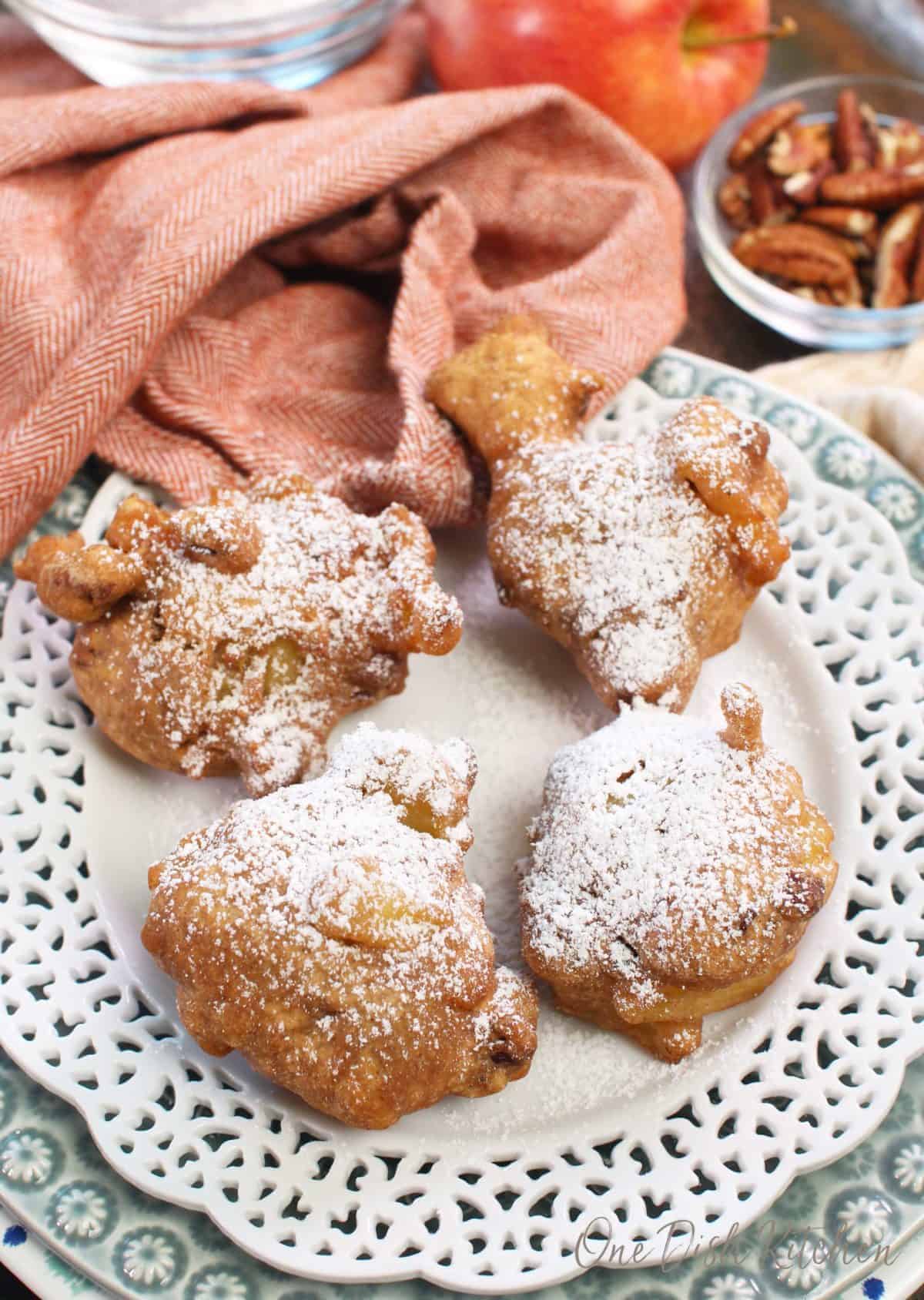 Four fritters on a white plate next to pecans and an apple.