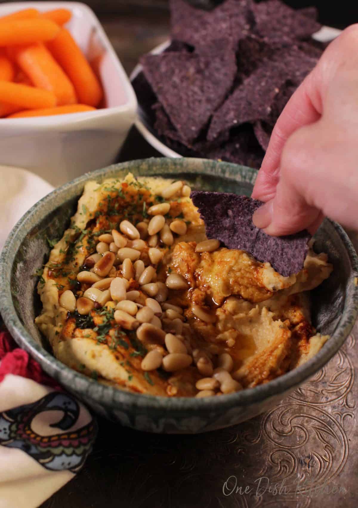 A blue corn chip scooping out hummus from a bowl that is topped with toasted pine nuts, olive oil, and paprika next to a bowl of blue corn tortilla chips and a small bowl of baby carrots