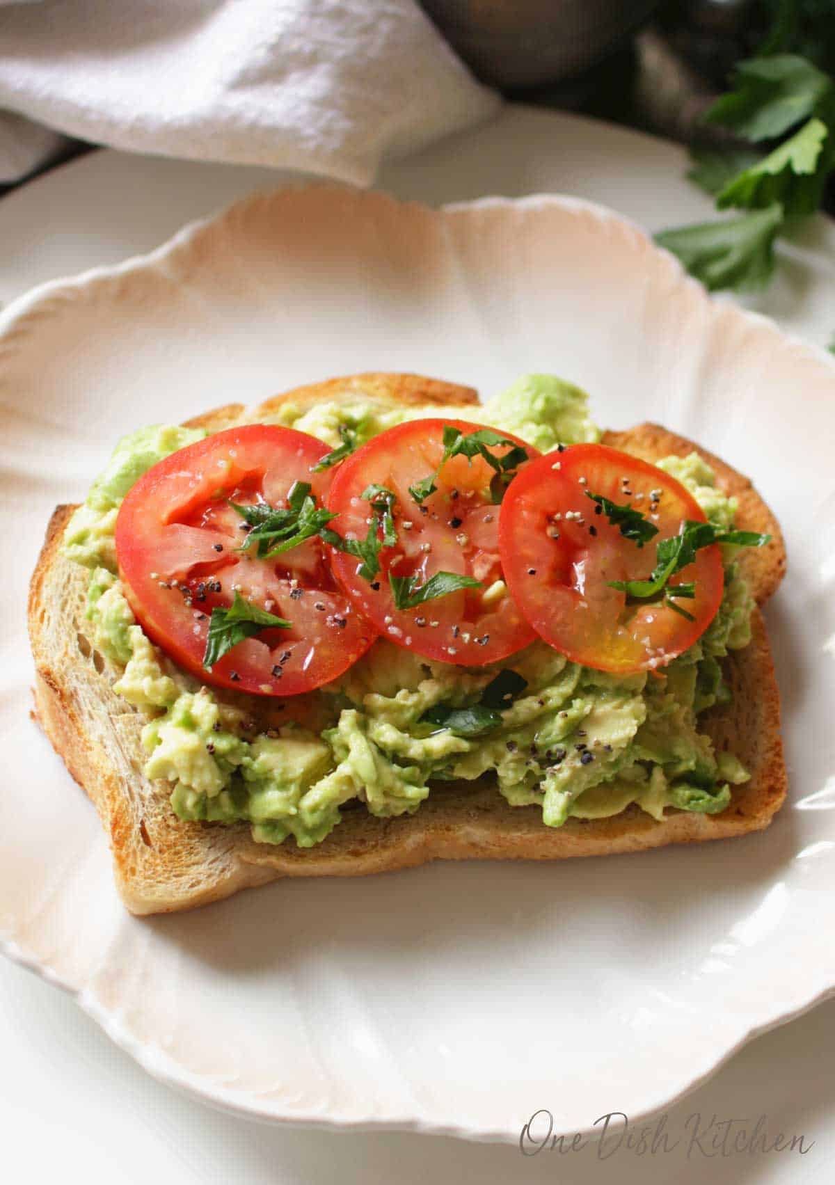 Avocado toast topped with three tomato slices, salt, pepper, and basil on a plate