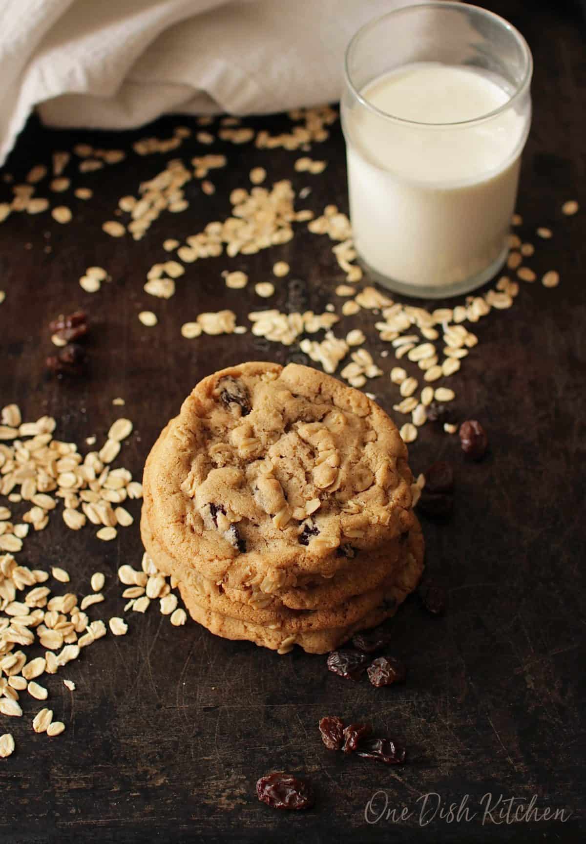 A stack of four oatmeal cookies on a dark surface with scattered oats and raisins and a glass of milk in the background