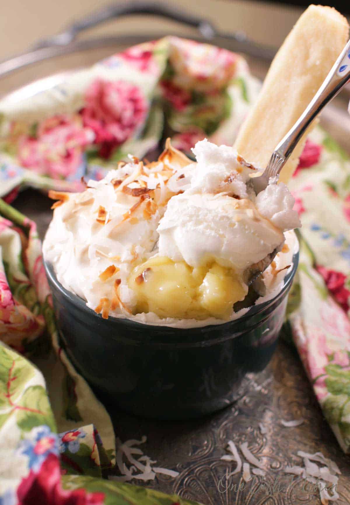 A spoonful of single serving coconut cream pie in a ramekin topped with coconut shavings next to a floral cloth napkin