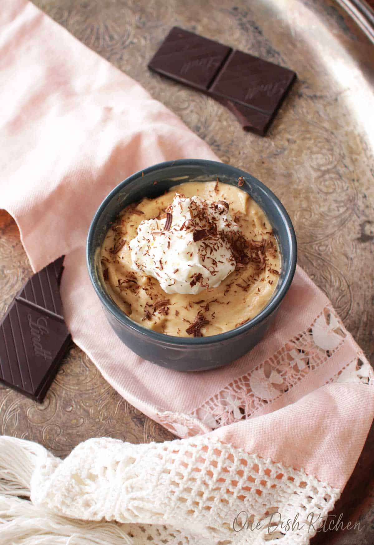 An overhead view of a small peanut butter pie in a blue bowl topped with whipped cream and chocolate shavings on a metal tray with a pink cloth napkin and four chocolate bar pieces