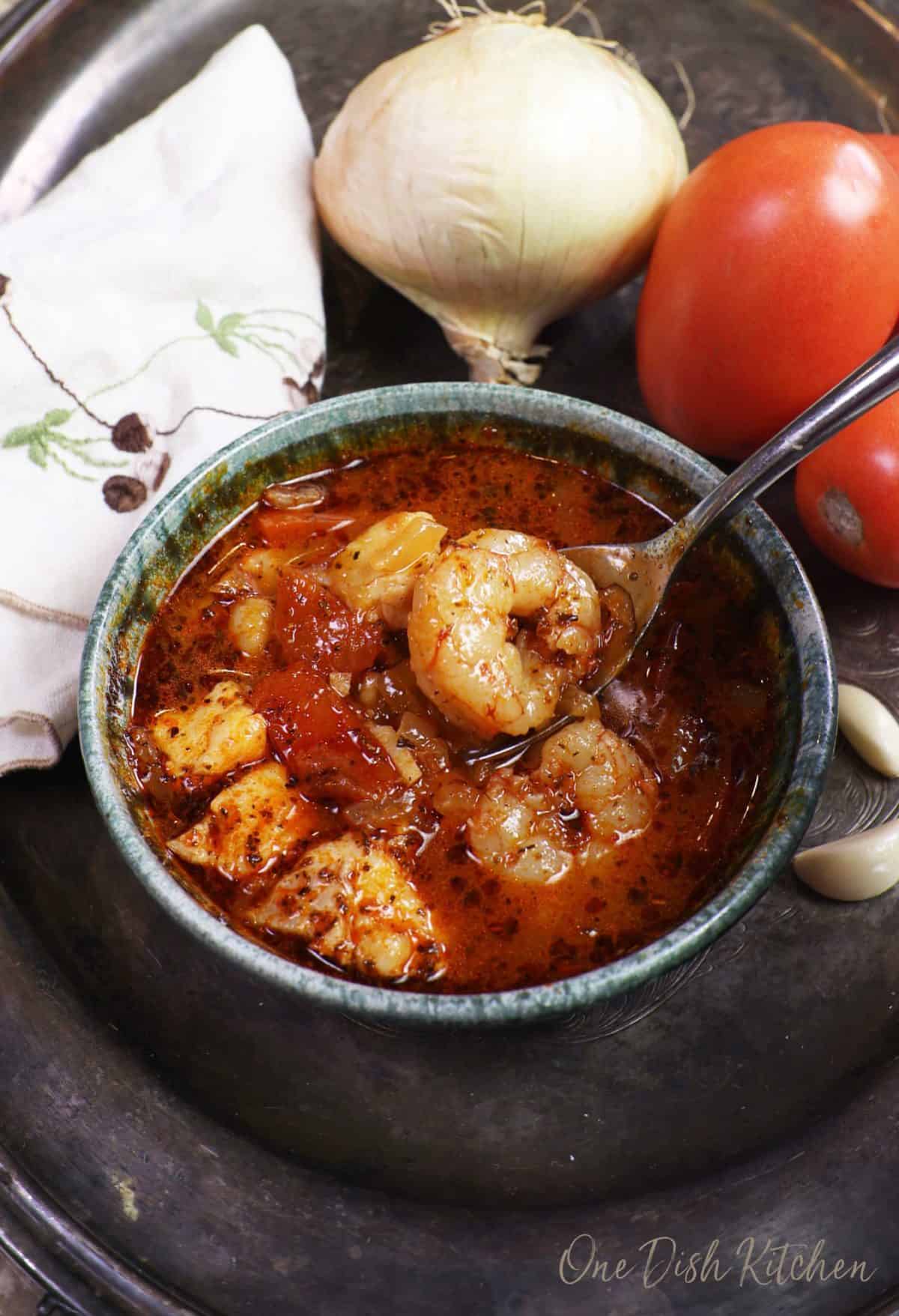 a blue bowl filled with cioppino with a large shrimp on a spoon above the dish.