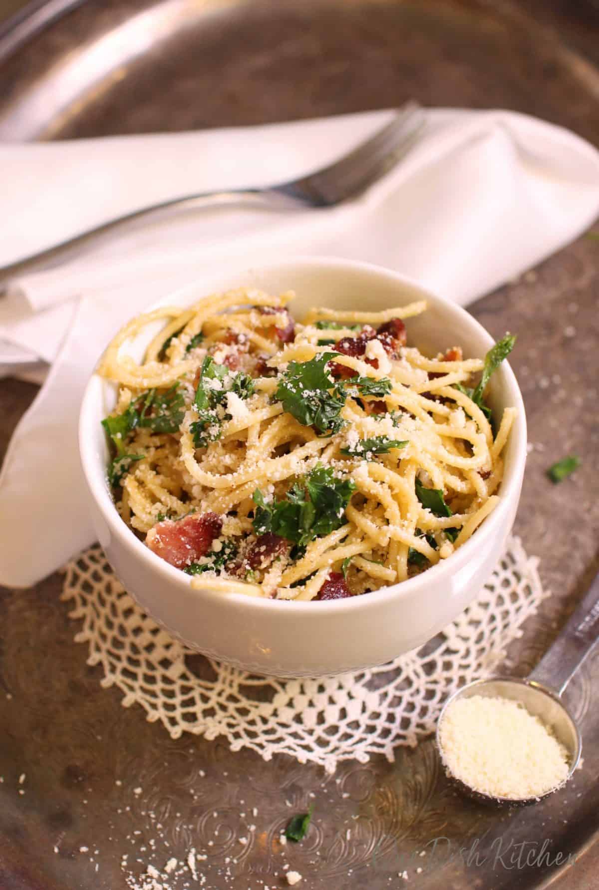 An overhead view of a bowl of pasta carbonara topped with chopped parsley and parmesan cheese next to a white cloth napkin, a fork, and measuring spoon of grated parmesan cheese all on a metal tray