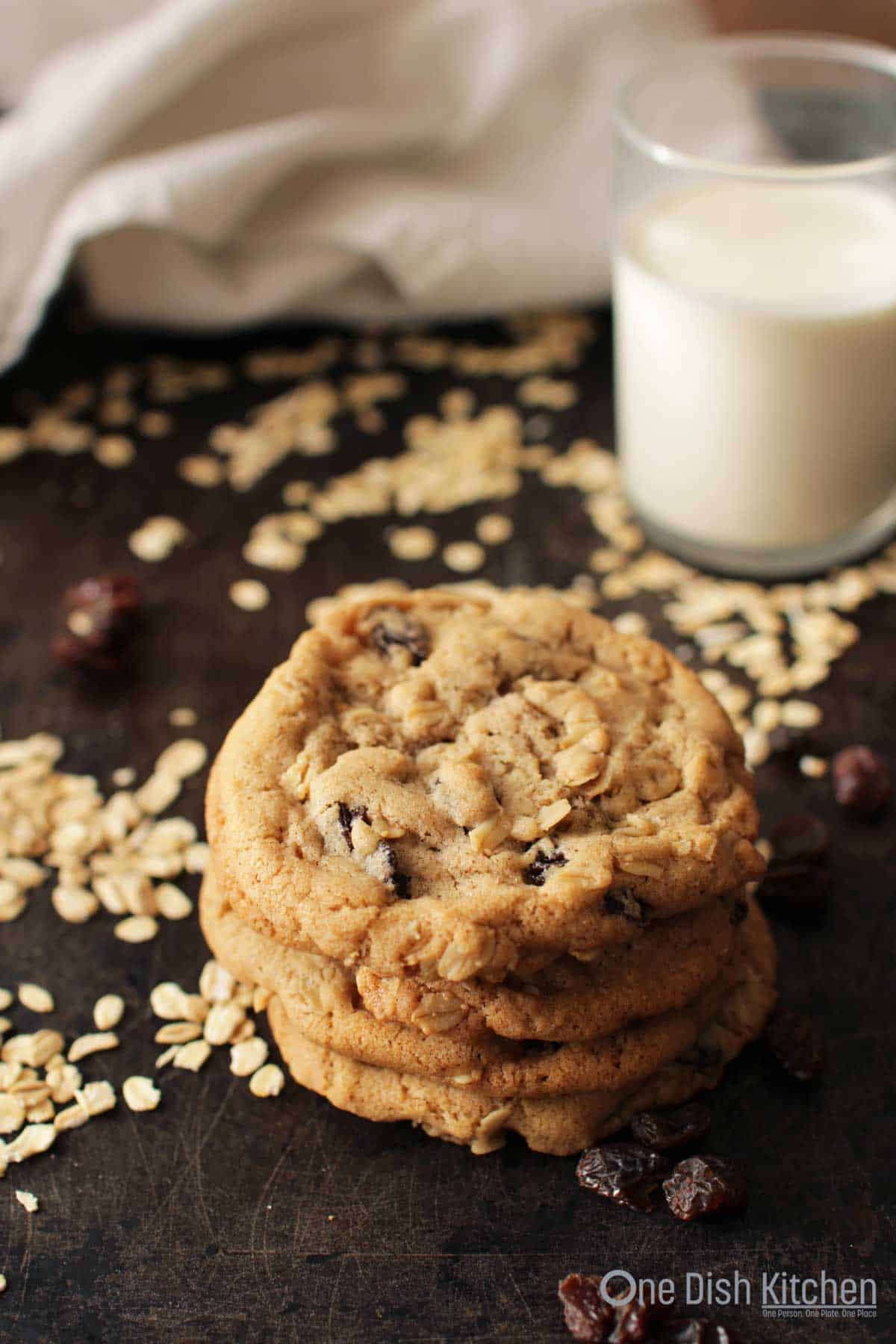 A stack of four oatmeal cookies on a dark surface with scattered oats and raisins and a glass of milk in the background