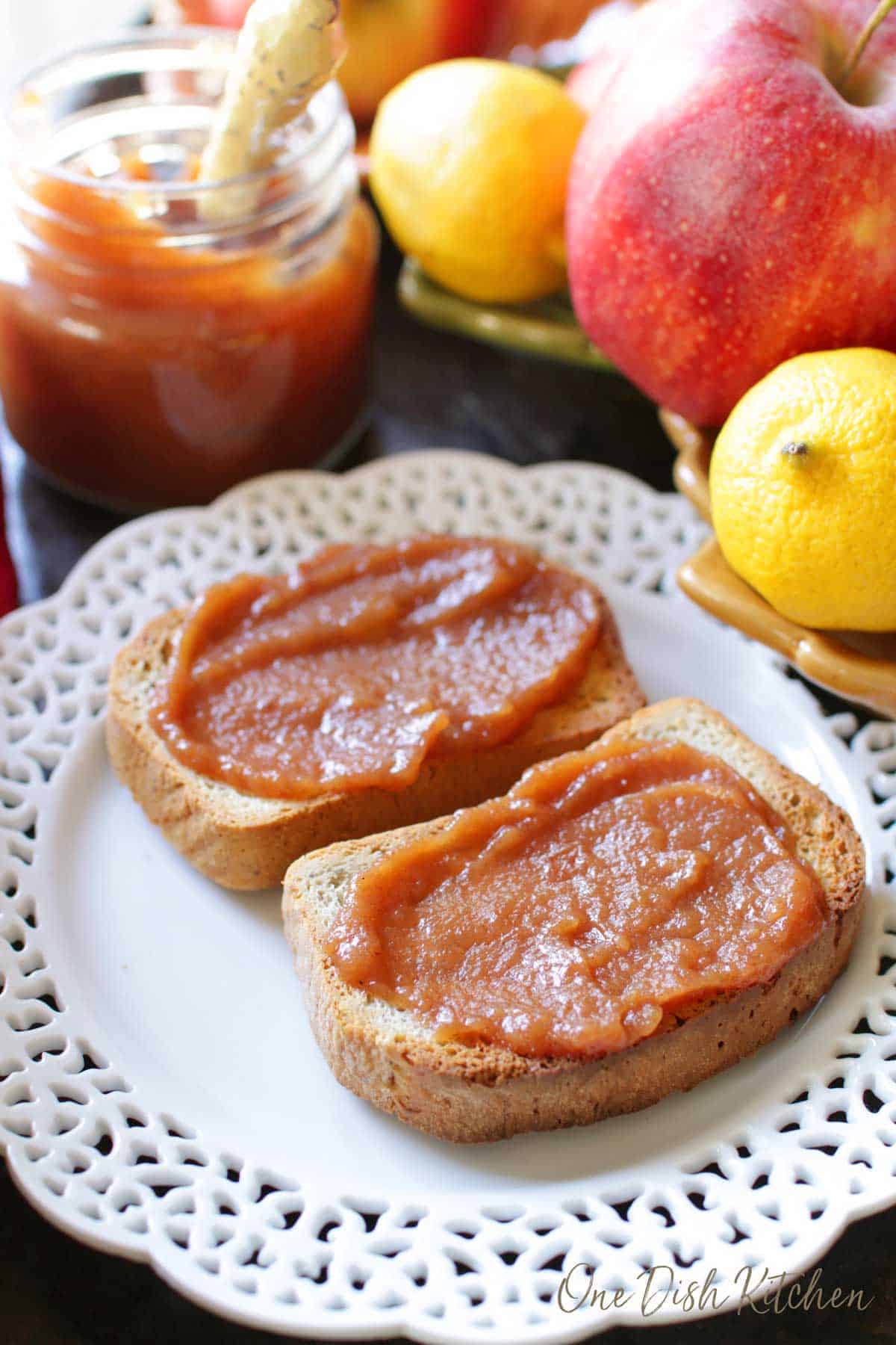 Apple butter spread on two slices of toast plated next to a small jar of apple butter with a knife and a bowl of apples and lemons