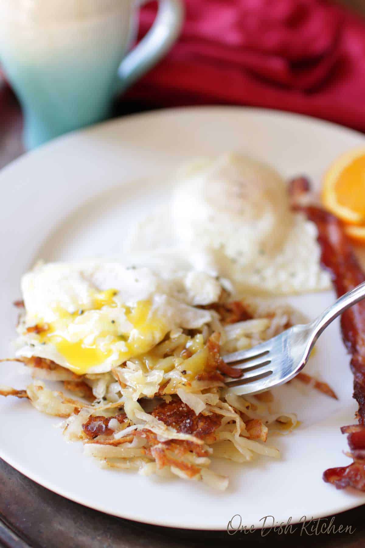 A forkful of hash browns and runny eggs from a plate with slices of bacon and orange slices