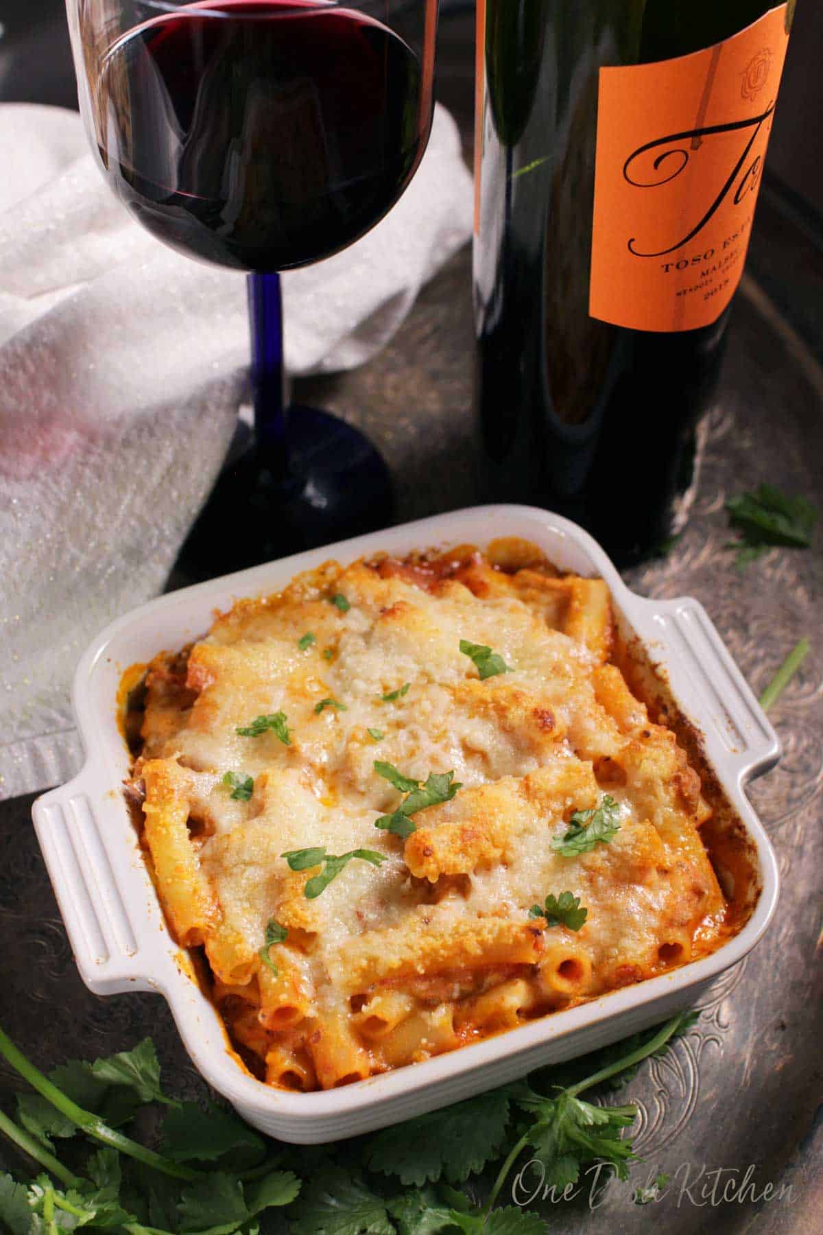 An overhead view of a small baked ziti casserole baked in a small baking dish on a metal tray with a glass and bottle of red wine and parsley