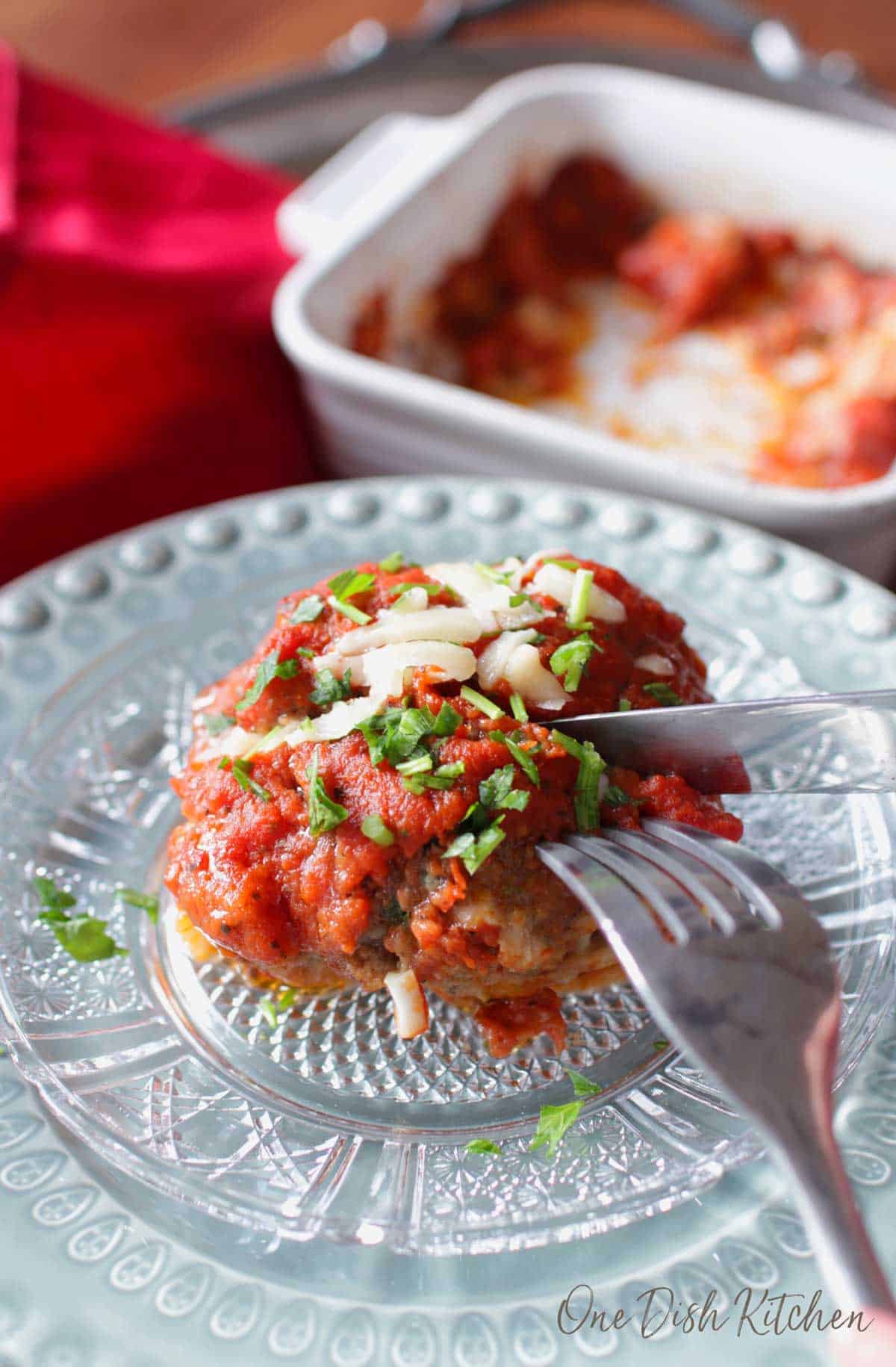 A fork and knife cutting into a meatball topped with tomato sauce and melted parmesan cheese