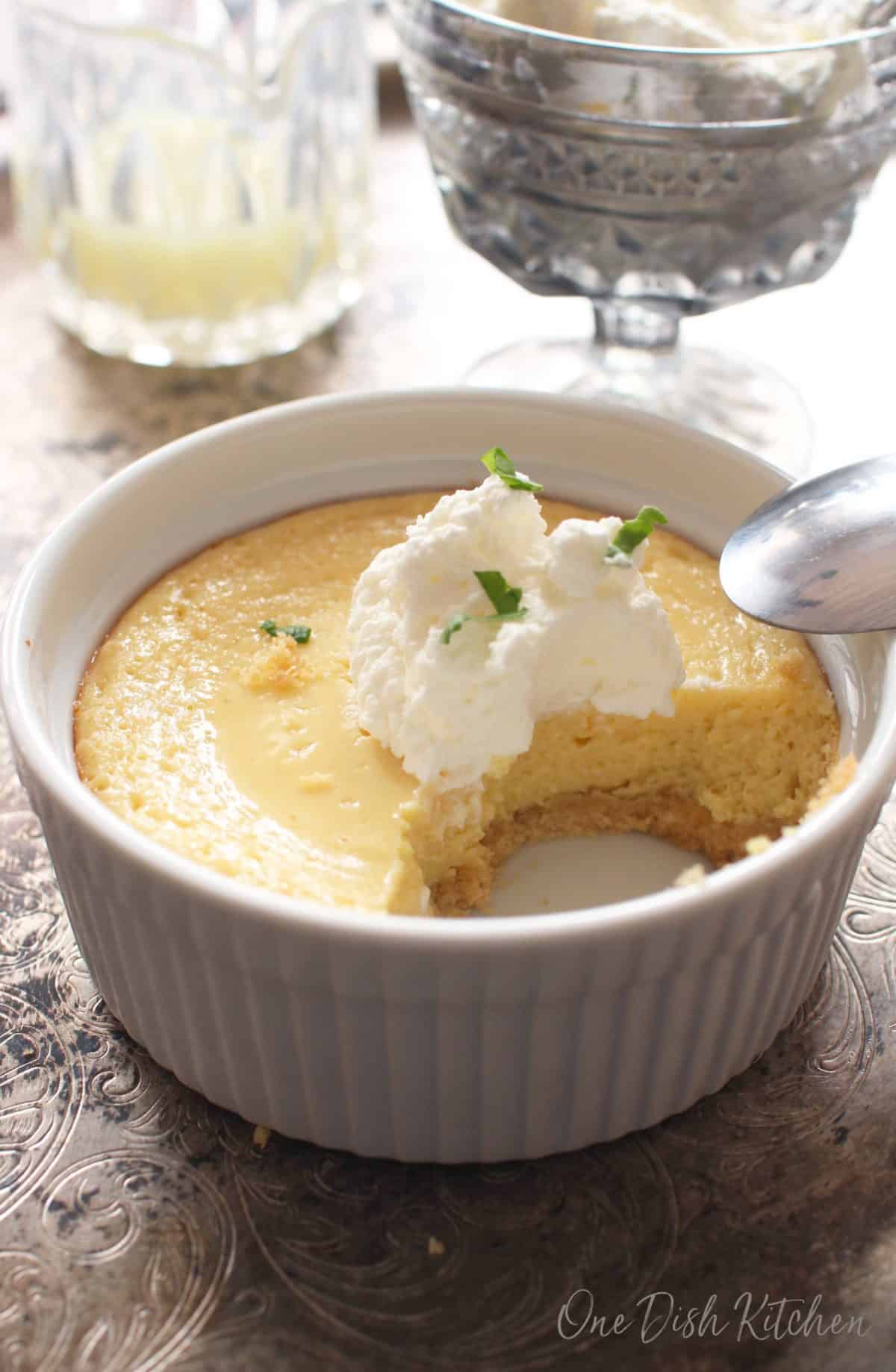 A partially eaten key lime pie topped with whipped cream with a spoon resting on the edge of the dish with a jar of lime juice and a dish of whipped cream in the background