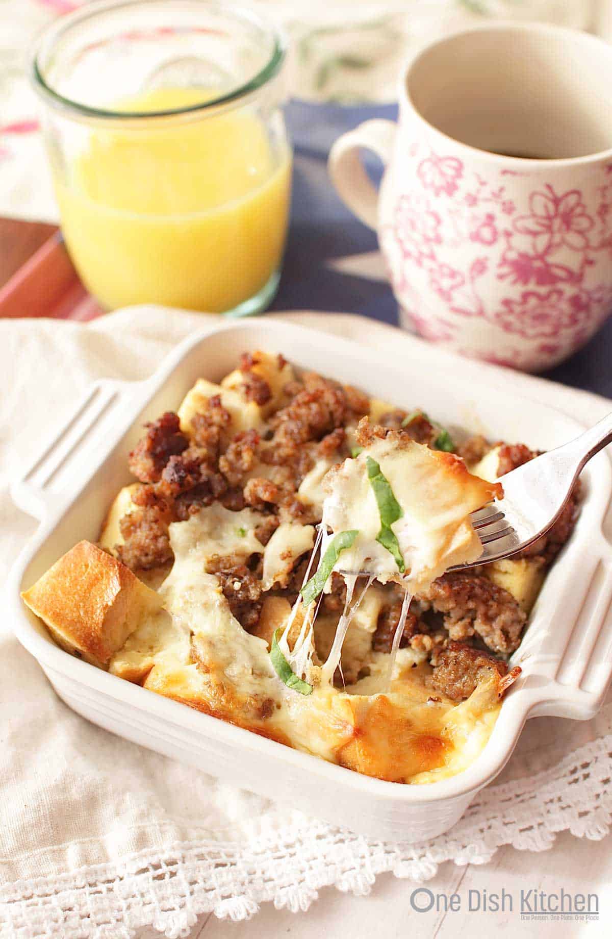 A forkful of breakfast casserole from a small baking dish next to a glass of orange juice and a coffee mug