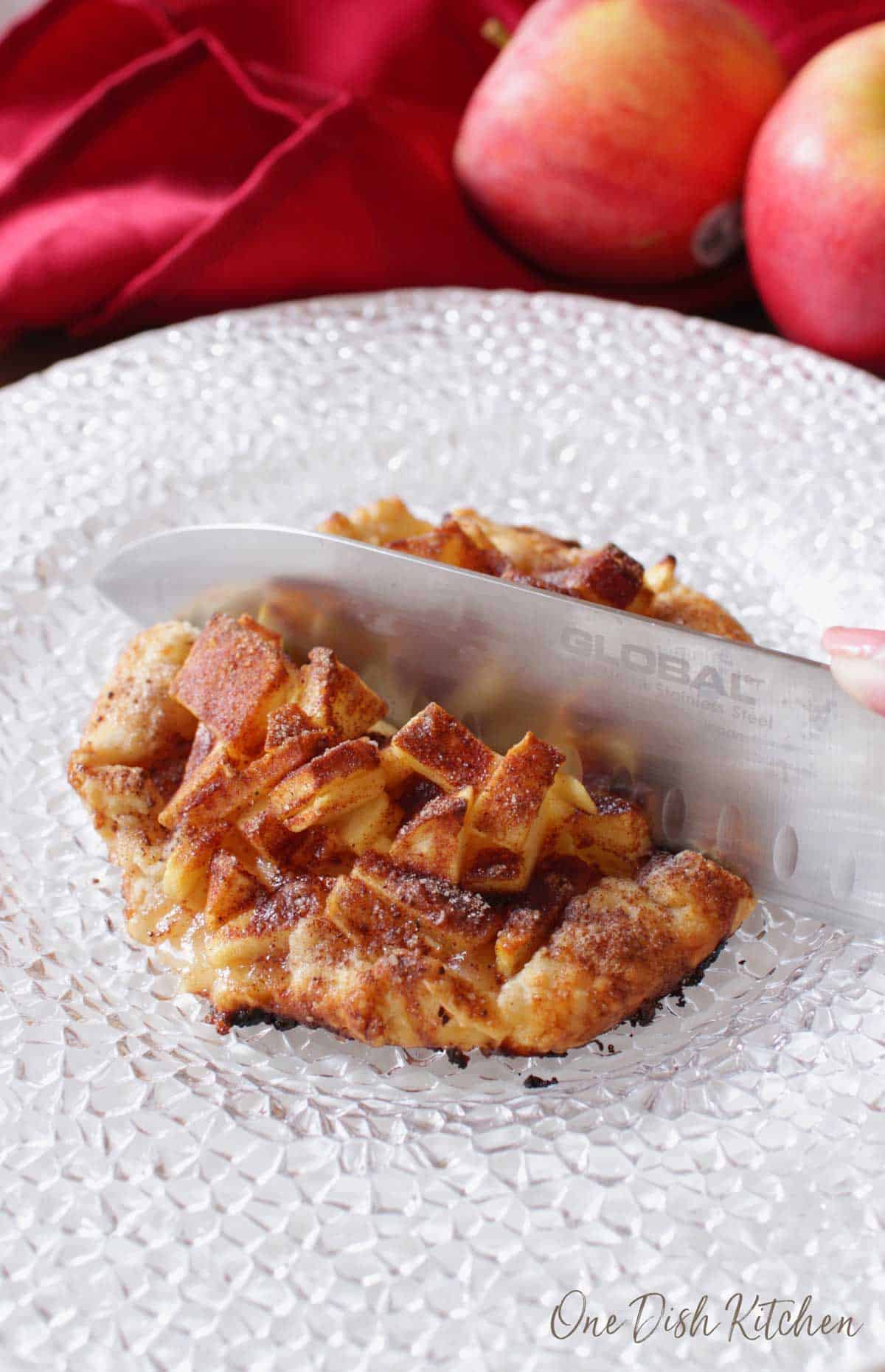 A knife cutting into an apple galette topped with cinnamon and sugar plated with apples and a red cloth napkin in the background