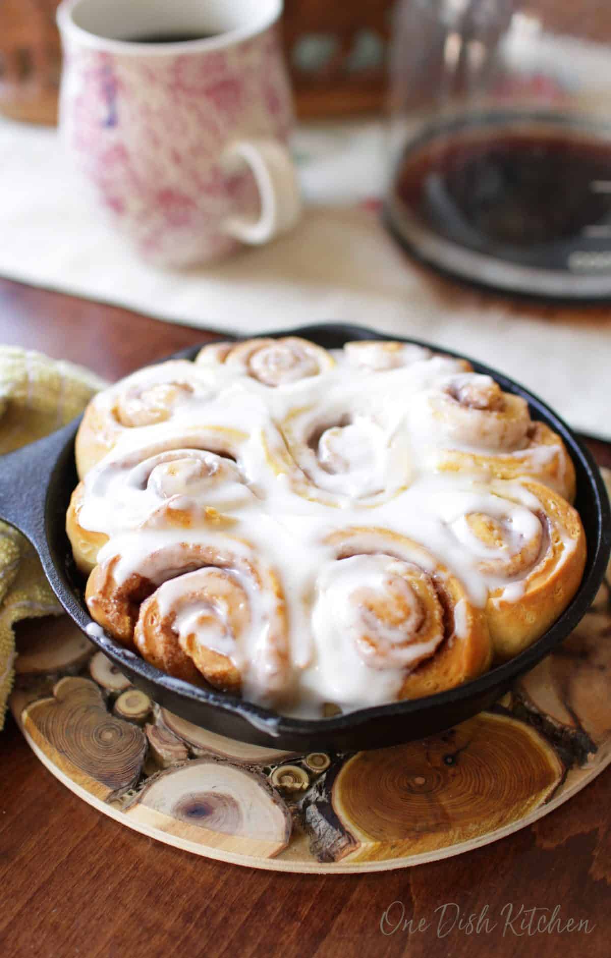 A small skillet of homemade cinnamon rolls topped with frosting on a wooden trivet