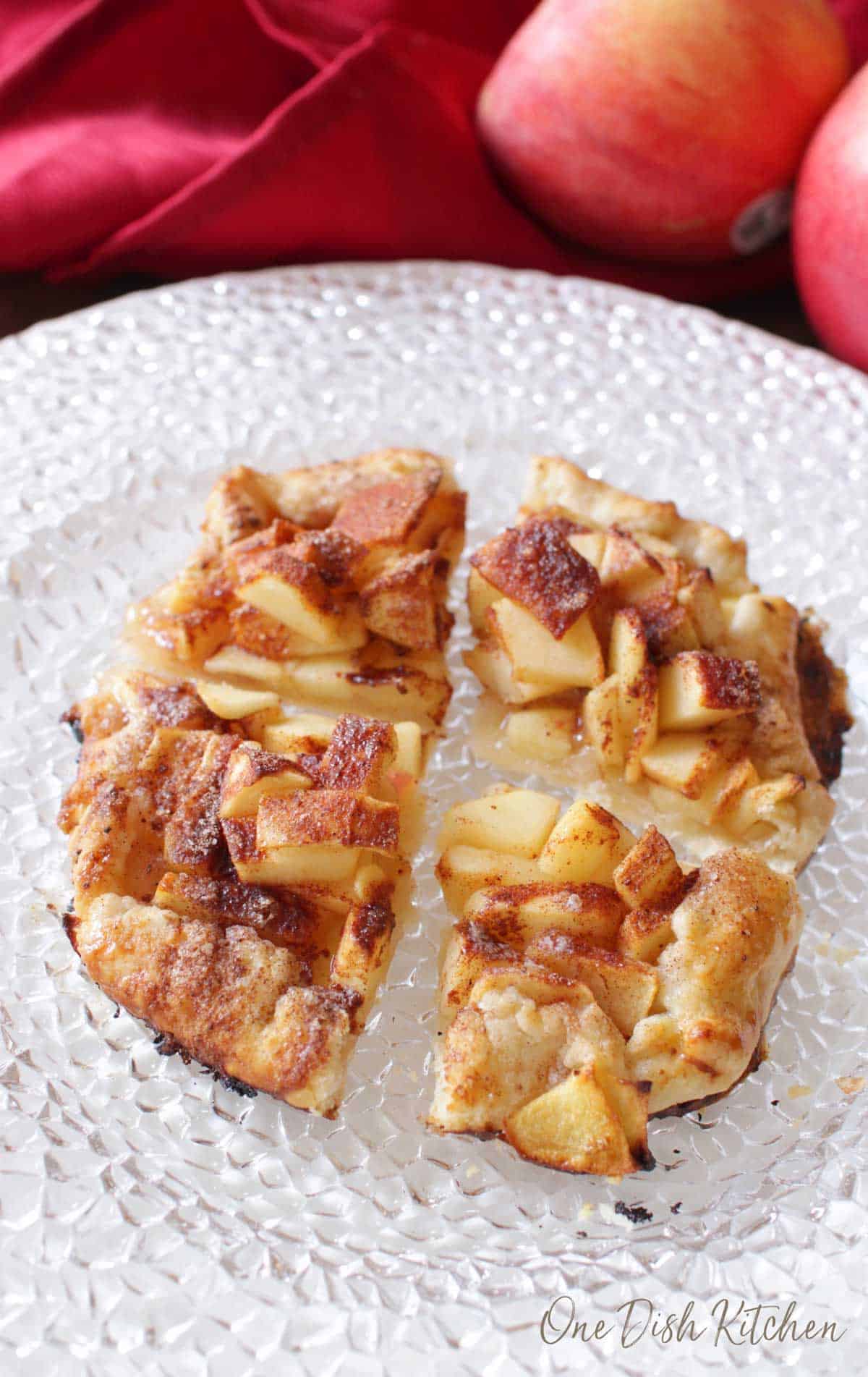 An apple galette topped with cinnamon and sugar cut into four slices plated with apples and a red cloth napkin in the background