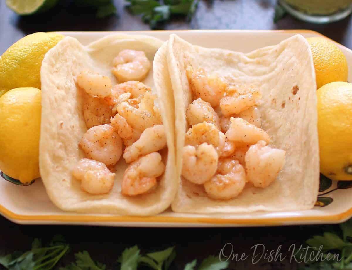 Two flour tortillas filled with shrimp on a plate surrounded by four whole lemons
