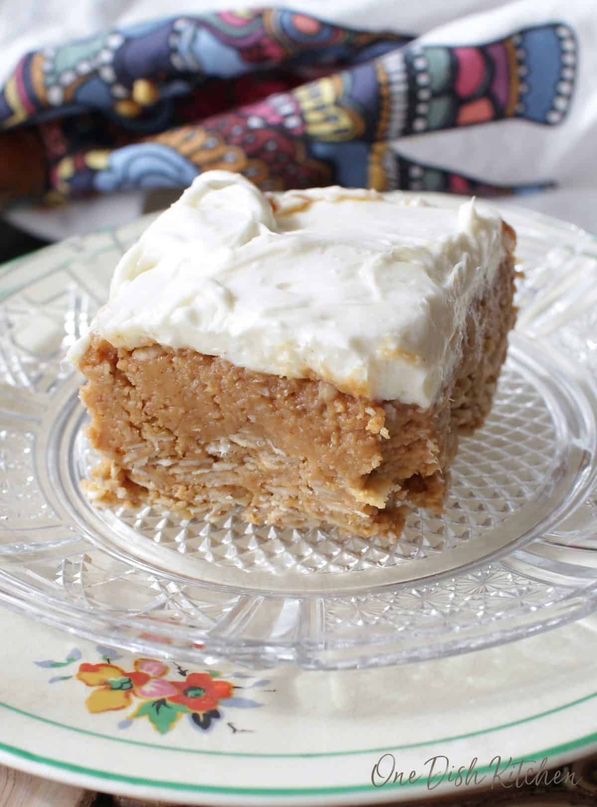 a square peanut butter bar with a white frosting on a clear plate next to a colorful napkin.