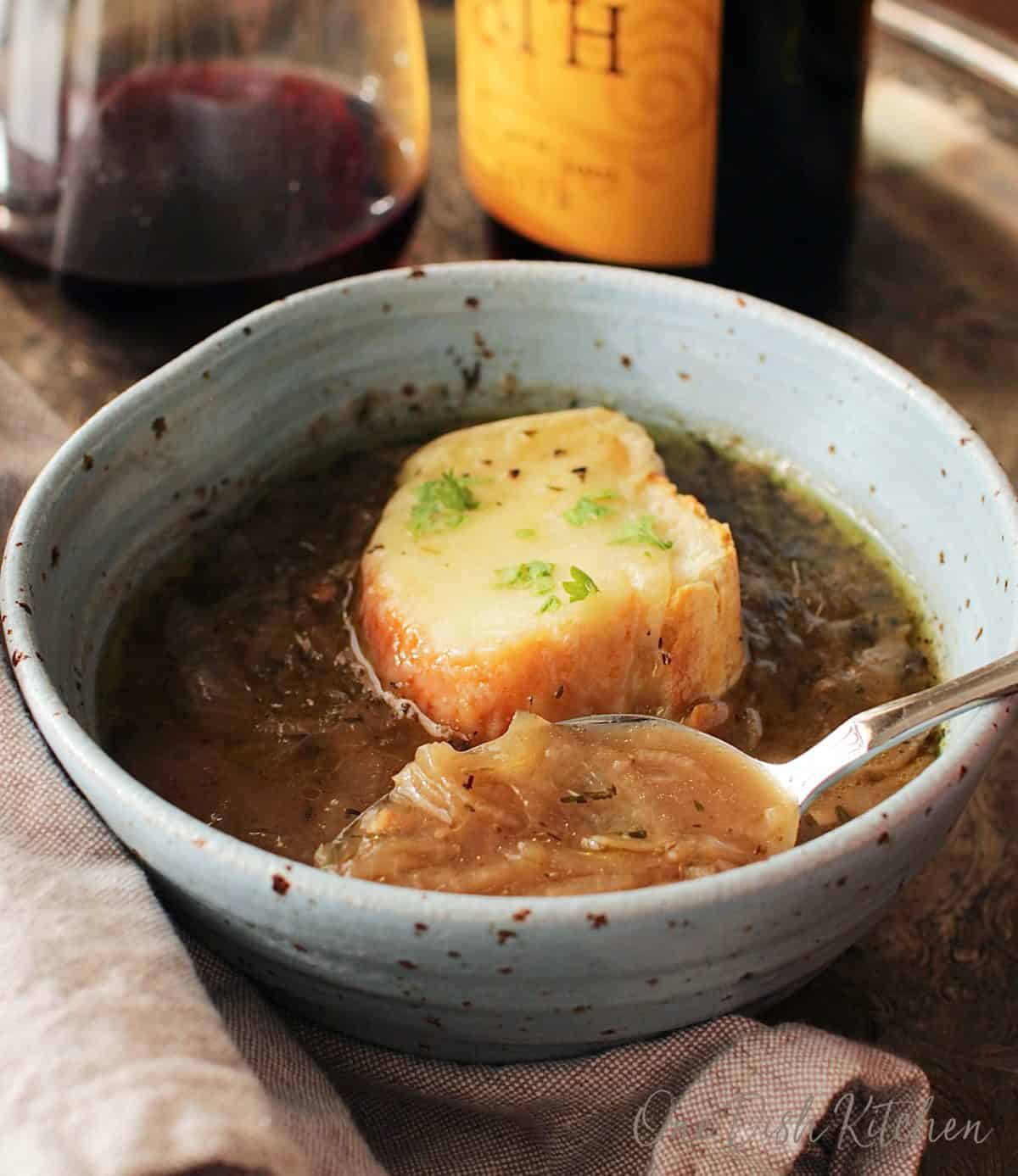 A spoonful of french onion soup from a bowl on a metal tray with a glass and bottle of red wine
