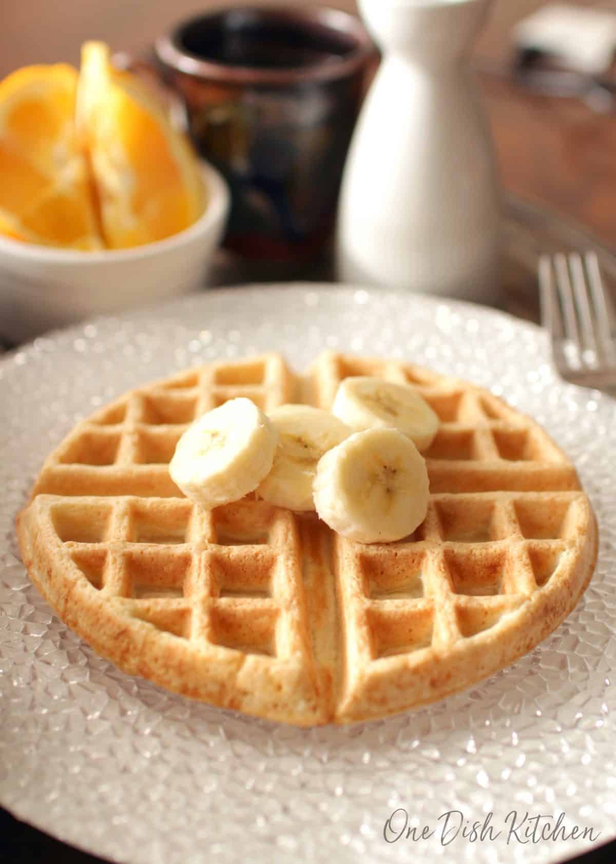 A waffle topped with banana slices on a large plate with a mug of coffee and orange slices in the background