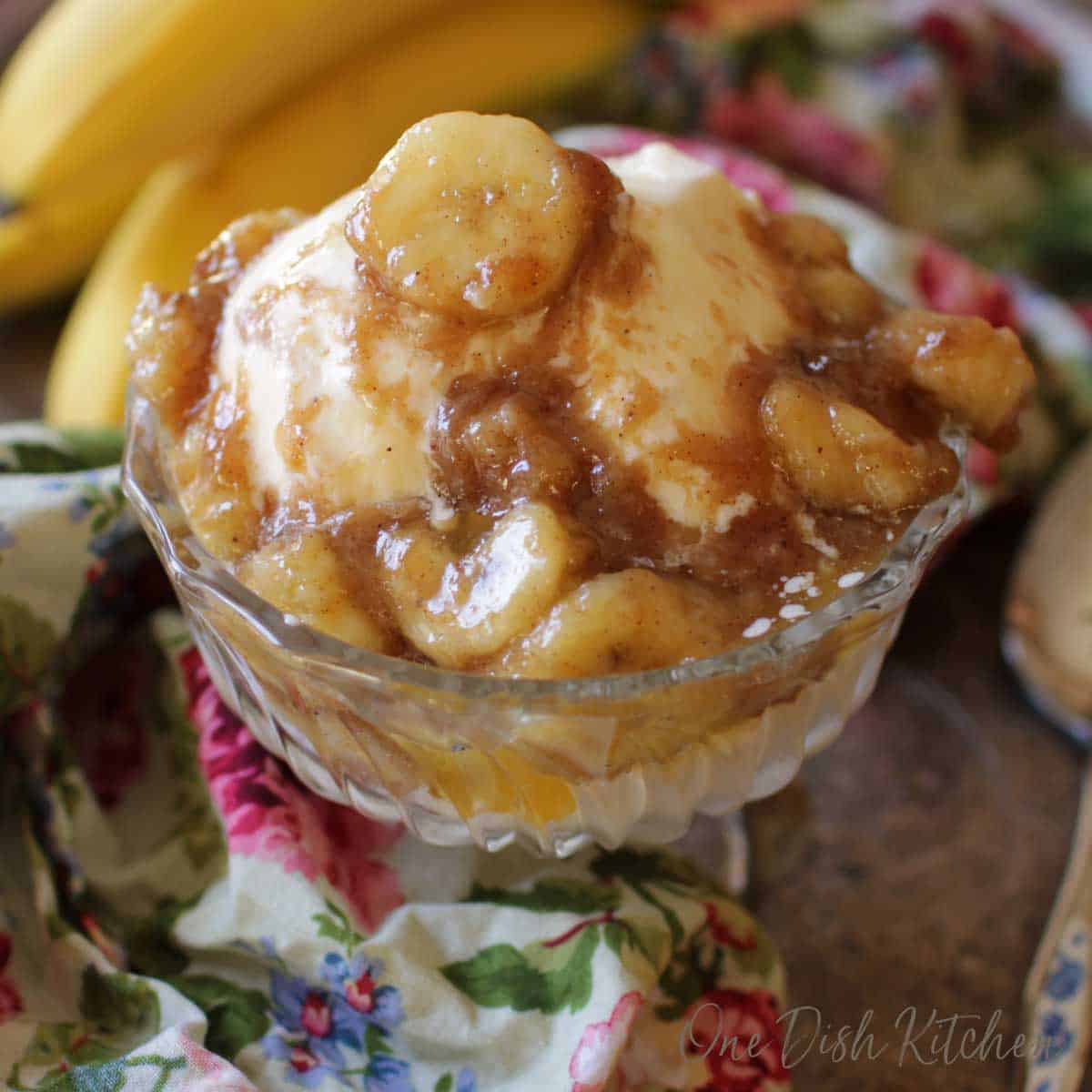 A dessert glass of vanilla ice cream that is topped with bananas mixed with brown sugar, butter and vanilla on a metal tray with a floral cloth napkin and a bunch of bananas