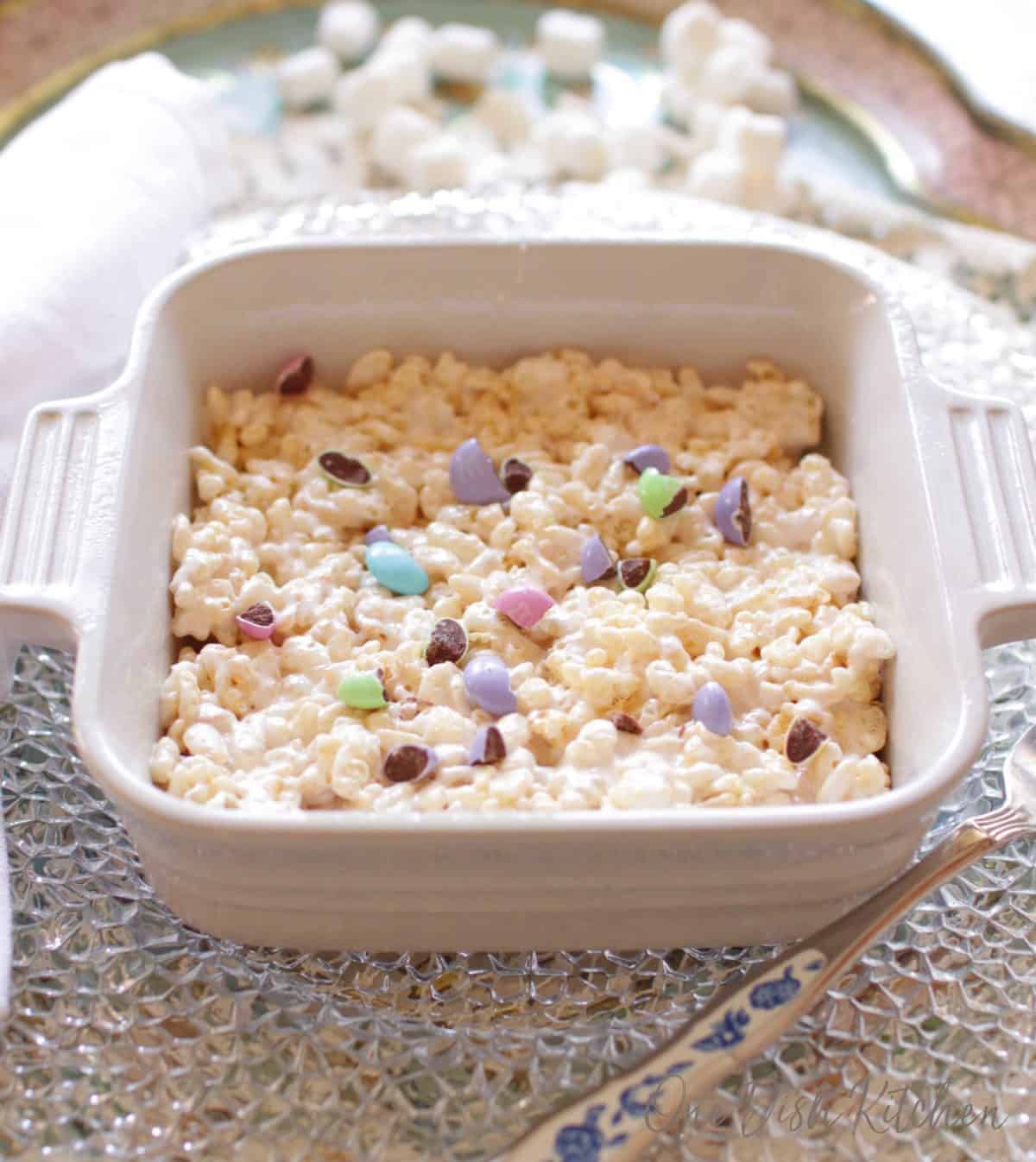 A giant rice krispies treat topped with pastel colored chocolate candies in a small baking dish next to a fork