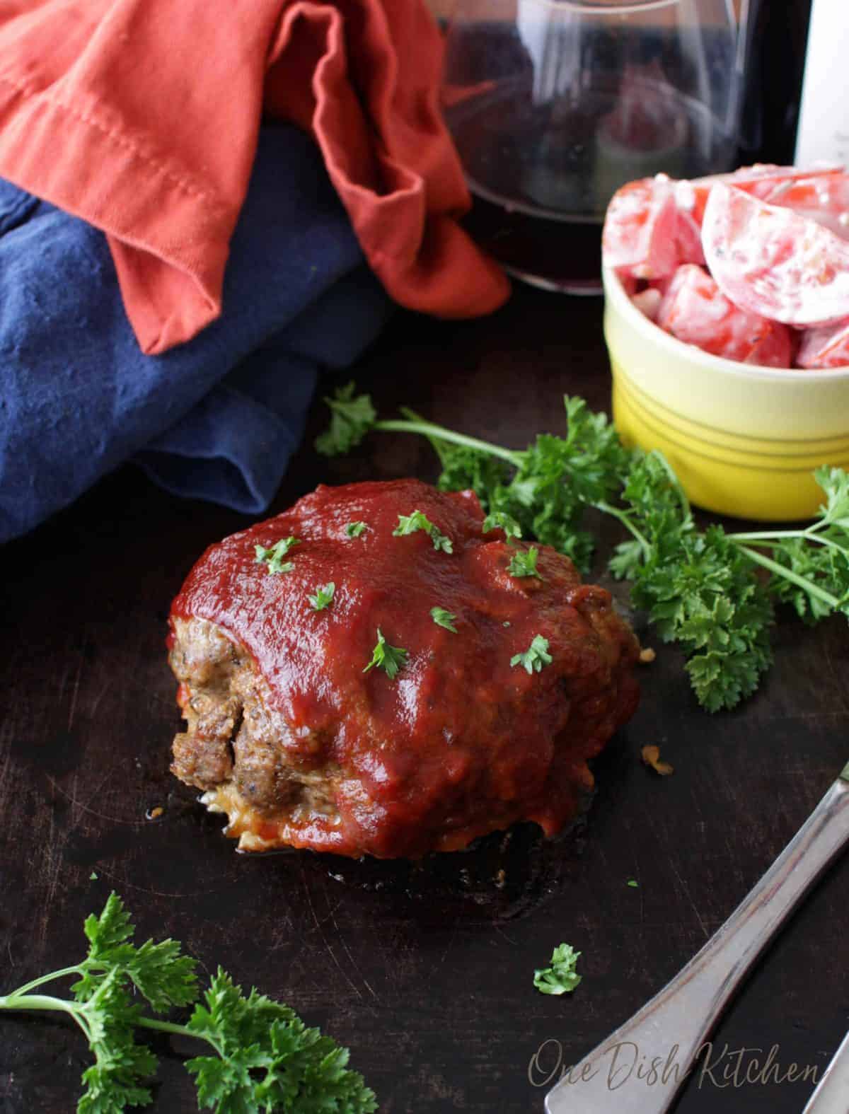 A single serving of meatloaf on a dark wooden tray with a glass of red wine and a small bowl of tomato slices with ranch dressing