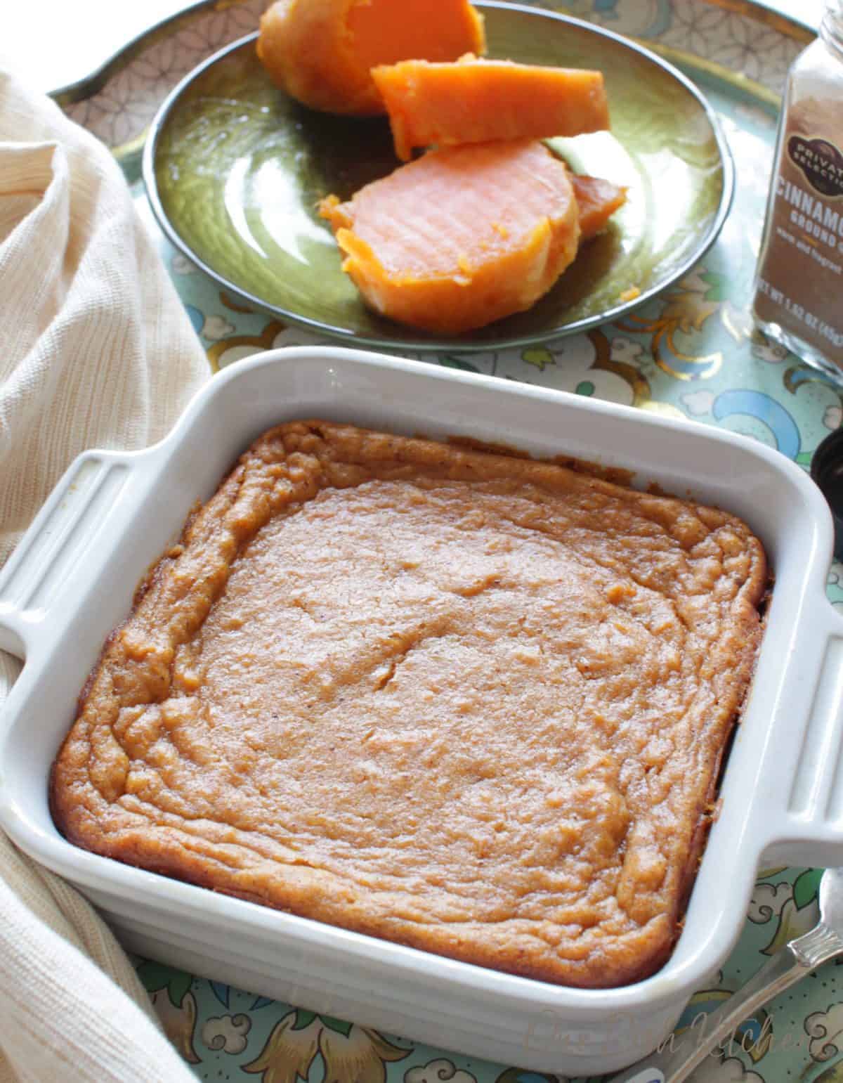 A sweet potato pie in a small baking dish on a tray with a sliced up sweet potato on a plate behind it and a shaker of ground cinnamon