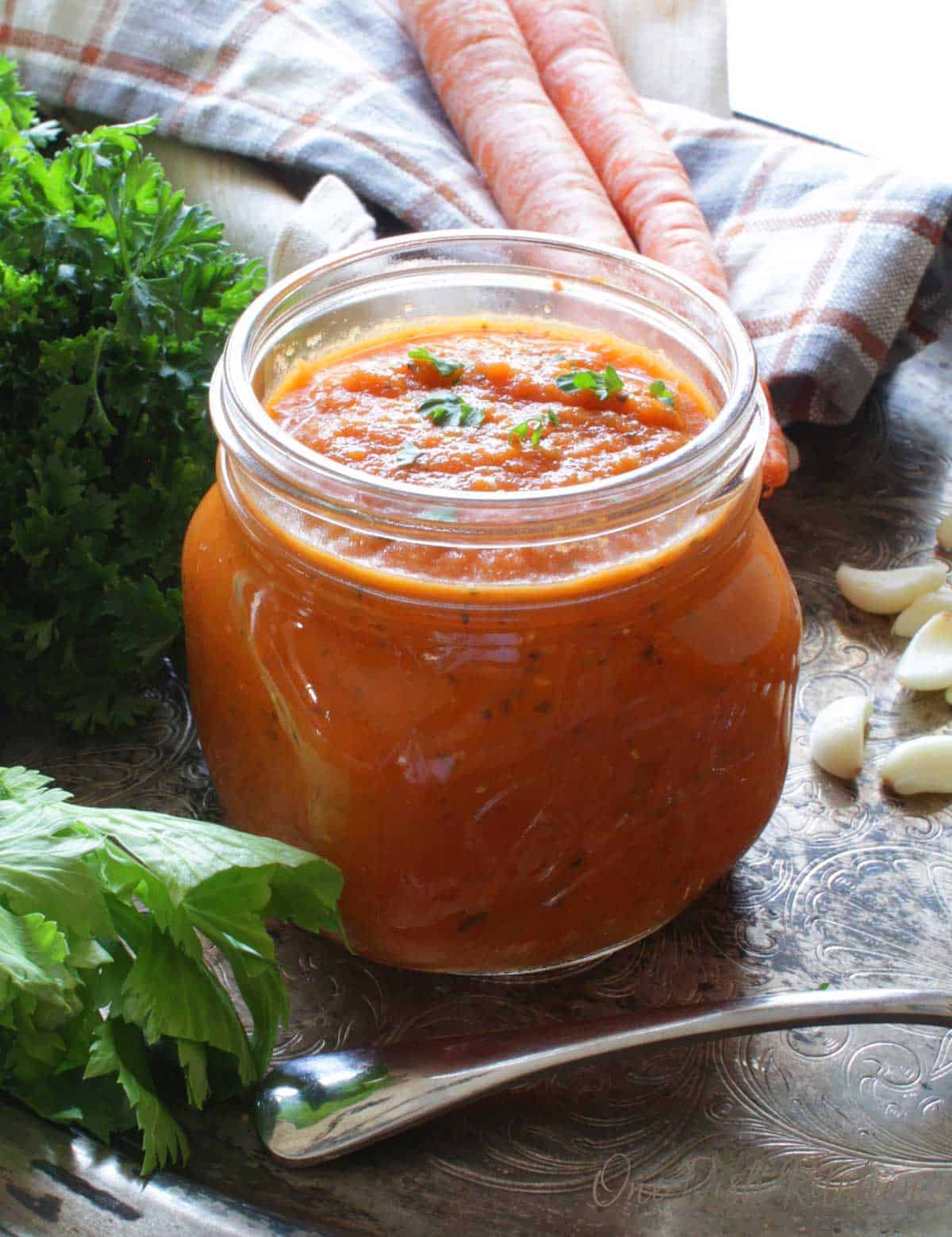 A small jar of homemade tomato sauce next to fresh carrots, parsley, and garlic cloves