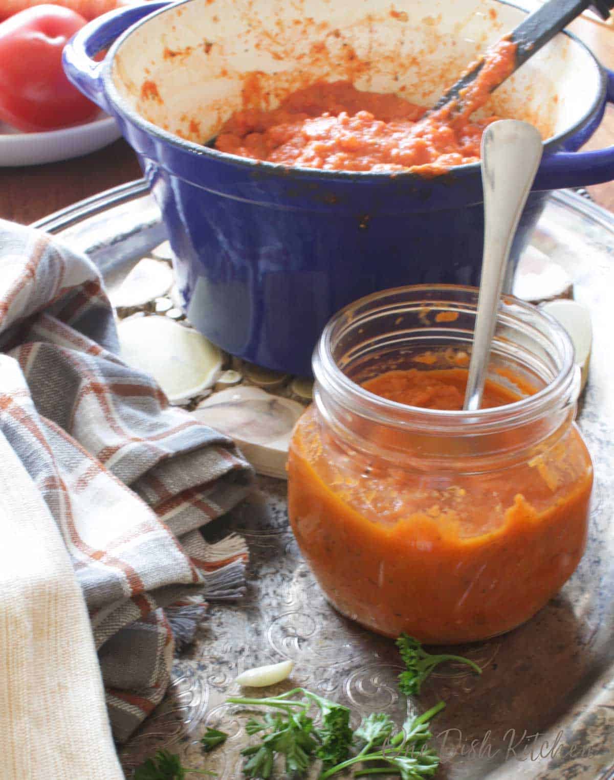 A small pot of tomato sauce next to a jar half filled with tomato sauce with a spoon all on a metal tray