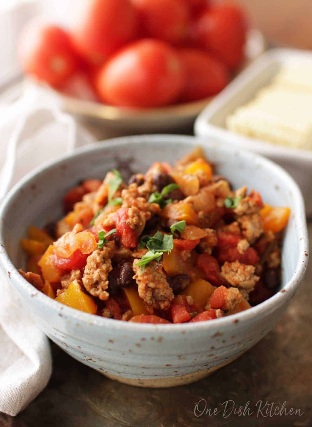 A bowl turkey chili in a blue bowl next to a plate of ripe red tomatoes.