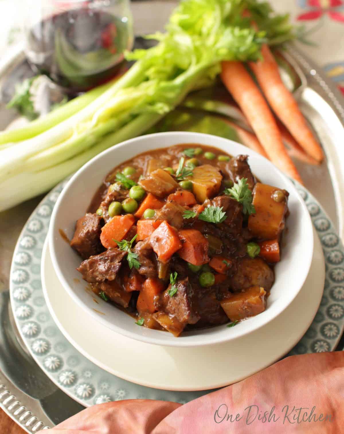 Overhead view of a bowl of beef stew next to two carrots, a stalk of celery, and a glass of red wine all on a metal tray