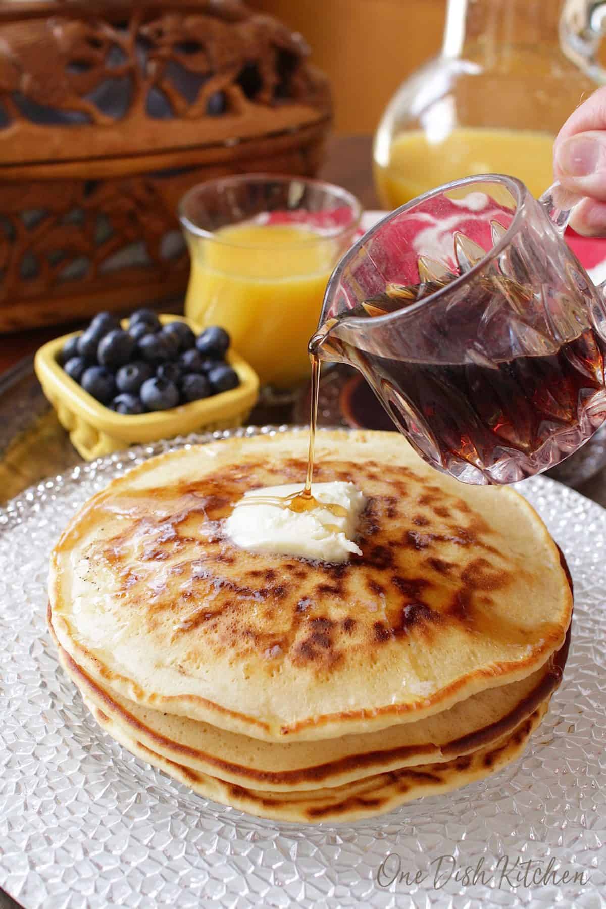 Pouring syrup over stack of pancakes on a plate topped with melting butter next to a small bowl of blueberries and a glass of orange juice