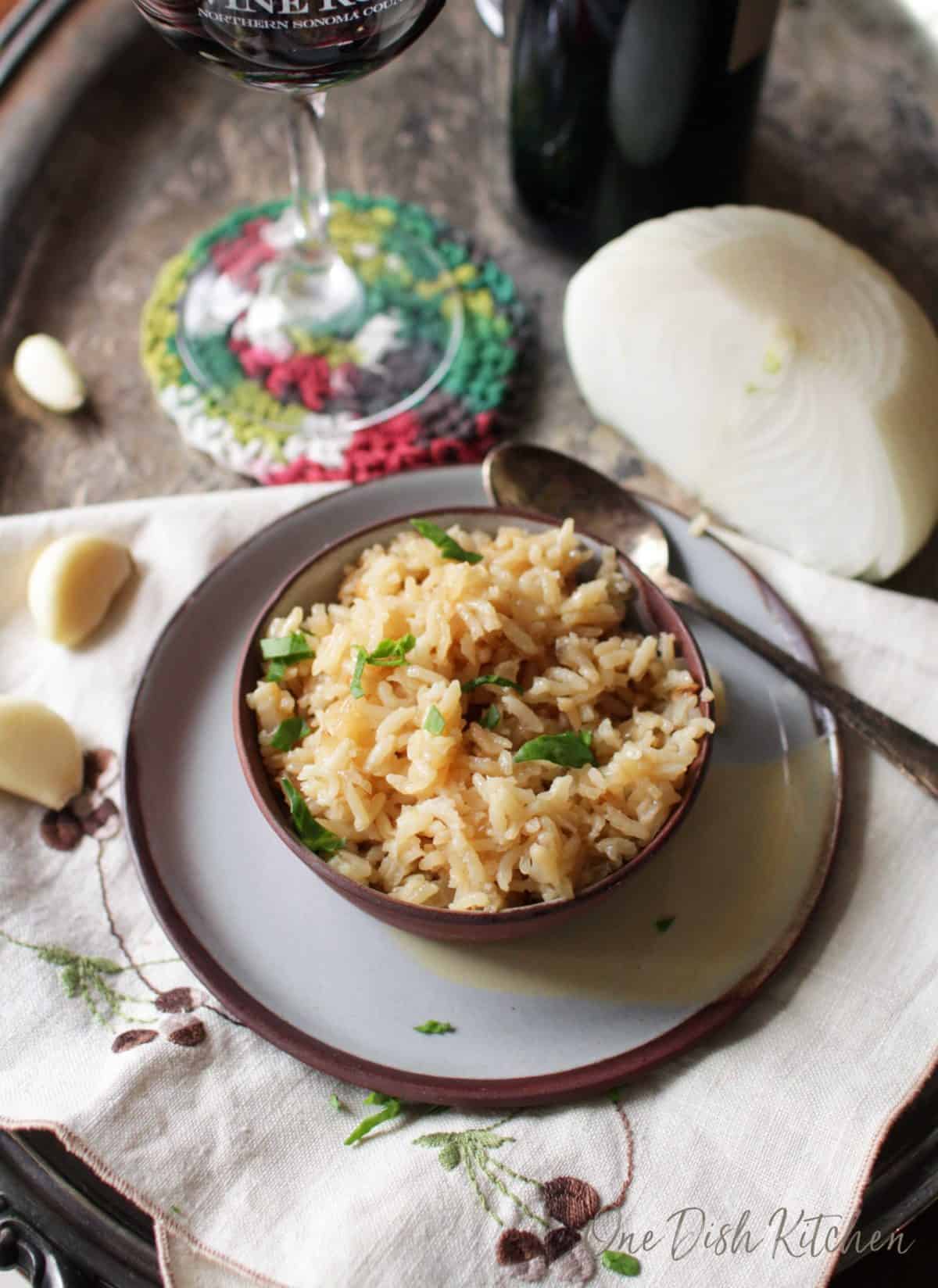 An overhead view of a bowl of rice pilaf next to a spoon, half an onion, a few garlic cloves, and a glass of red wine