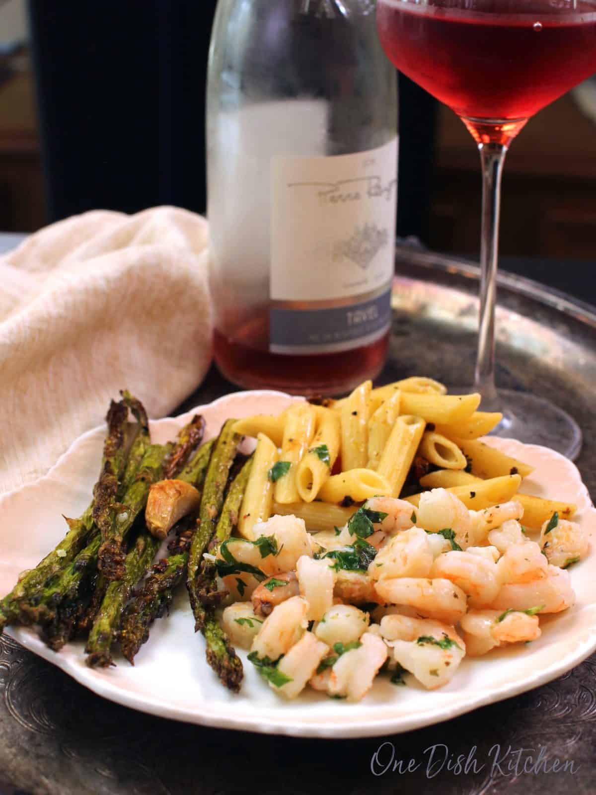 a white plate filled with shrimp, asparagus and penne pasta on a silver tray next to a glass of rosé and a bottle of wine in the background.