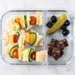 A lunch box with cheese sandwich skewers, pickles, and fruit.
