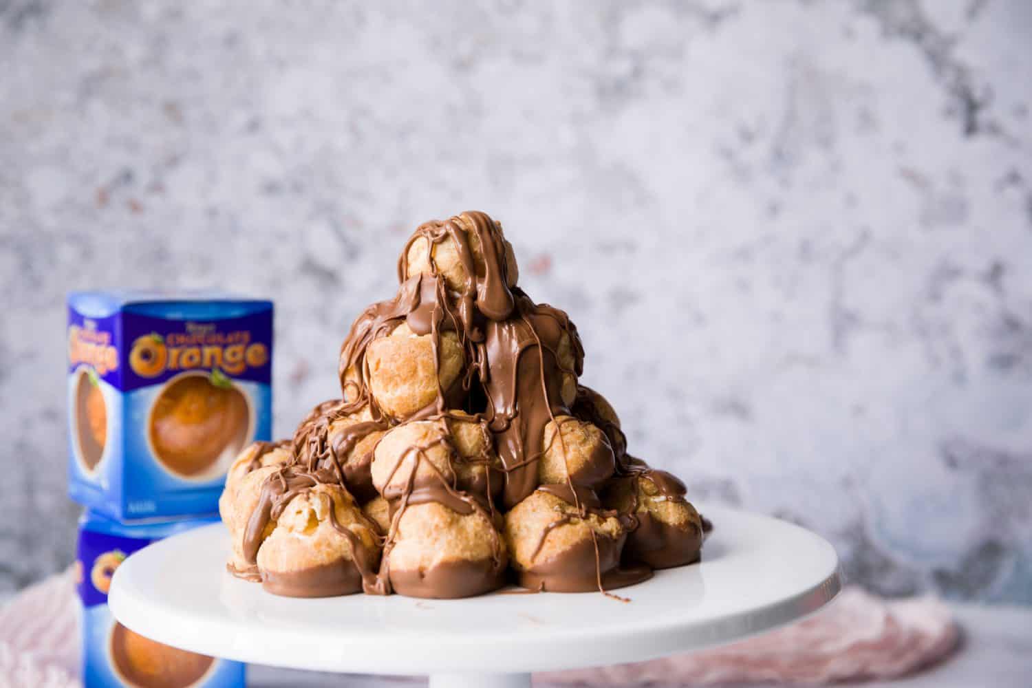 Um suporte de bolo branco com uma pilha de profiterole no topo que foi coberta com molho de chocolate. Há duas laranjas de chocolate Terry's no fundo.'s chocolate oranges in the background.