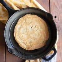 A lentil and chickpea flatbread sits inside a black cast iron pan, resting on a yellow tea towel atop a wooden table surface.