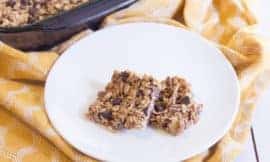 Two square choloate chip granola bars sit on a round white plate on top of a yellow dish towel.