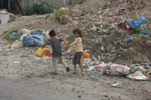 Two children holding plastic jugs walk away from the camera past a pile of rubble. One of the children is looking over her shoulder smiling.