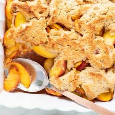 A baking dish filled with a golden brown peach cobbler with one scoop taken out.