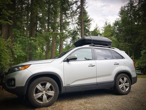 A silver SUV with a cargo box on the roof rests on a gravel driveway with pine trees in the background. One of the many gap year ideas for families is taking a road trip in a car and camping.
