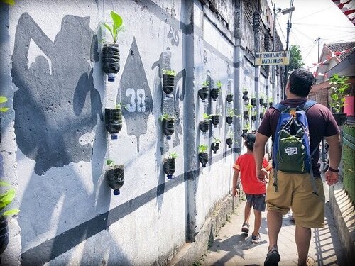 A man and a young boy walk through a small walkway in Yogyakarta, Indonesia, where travelers can learn Indonesian. To the left of them, they pass a wall decorated with plastic bottles that have been converted into planters, and paintings of shadow puppets on the wall.