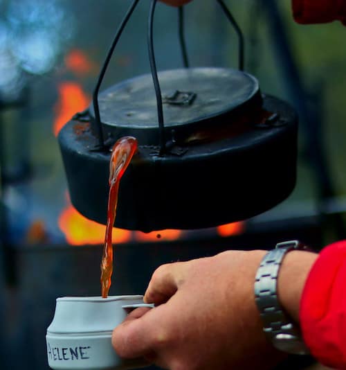 A person pours coffee from a beat-up kettle that has just come off an open fire
