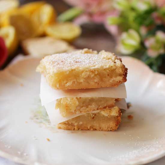 A stack of three lemon bars on a plate with lemon slices and pink flowers in the background