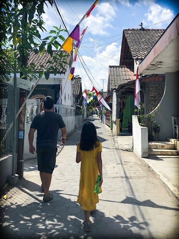 A father and daughter walk through a small neighborhood in Yogyakarta, Indonesia, where they can learn about Indonesian culture. The houses are very close together, and there are flags throughout the street.