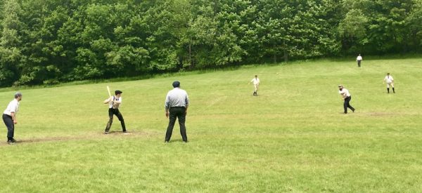 Men on a field in Cooperstown, NY playing baseball in historicl costumes and with historic gear and rules.