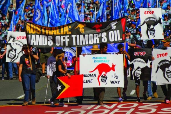 People marching with a large banner that reads AUSTRALIA: HANDS OFF TIMOR OIL. Another sign has a kangaroo graphic and the text MEDIAN LINE = 1/2 WAY. referencing the median line.