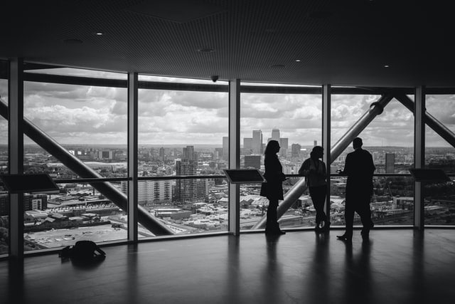 A black and white photo of three people meeting in a big room with glass windows overlooking a city. Photo by Charles Forerunner on Unsplash