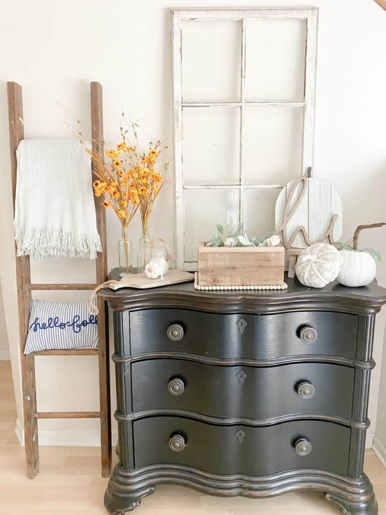 Entryway table vignette with muted fall decor. There is an old ladder with a blanket and pillow on it, an old window on the the table alongwith various fall decor items such as muted yellow flowers, white and white and gray patterned pumpkins, a wooden pumpkin sign with a wooden "hi" in front, and a wooden box with faux lambs ear and mini pumpkins inside.